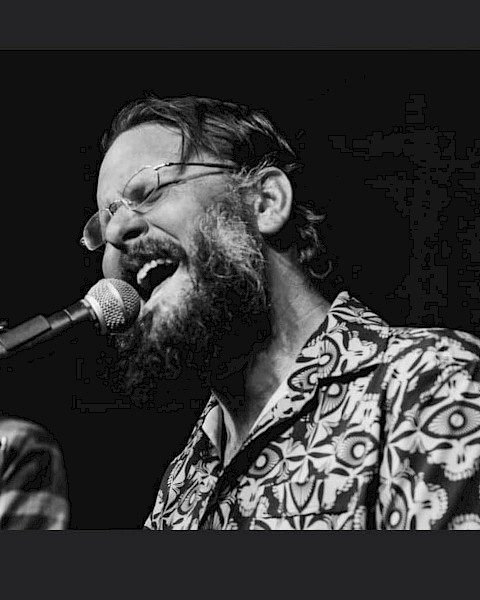 Evan Mullins wearing glasses and a patterned shirt passionately singing into a microphone on stage at Beats + Bites Brunch, live at Currents at Riverhouse Lodge in Bend Oregon on October 19