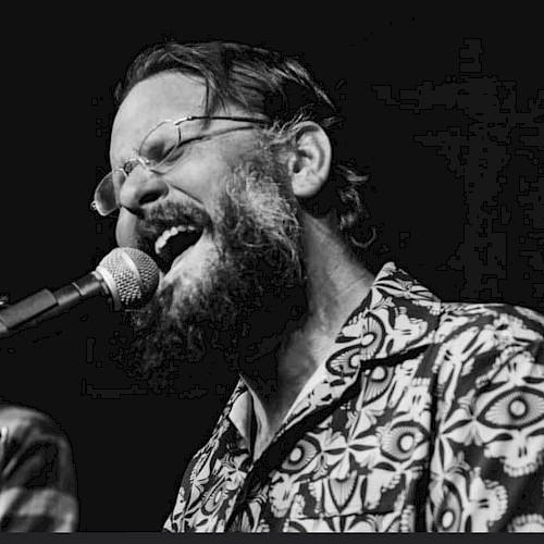Evan Mullins wearing glasses and a patterned shirt passionately singing into a microphone on stage at Beats + Bites Brunch, live at Currents at Riverhouse Lodge in Bend Oregon on October 19
