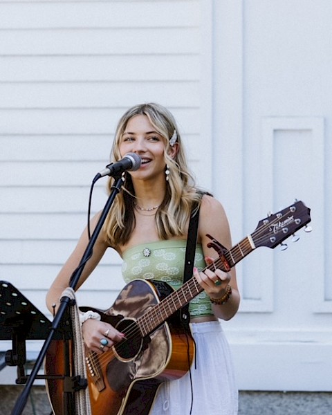 Samantha Hart playing guitar and singing into a microphone outdoors with a music stand and white building in the background, performing live at Currents at Riverhouse Lodge in Bend Oregon on October 26, 2025