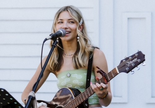 Samantha Hart playing guitar and singing into a microphone outdoors with a music stand and white building in the background, performing live at Currents at Riverhouse Lodge in Bend Oregon on October 26, 2025
