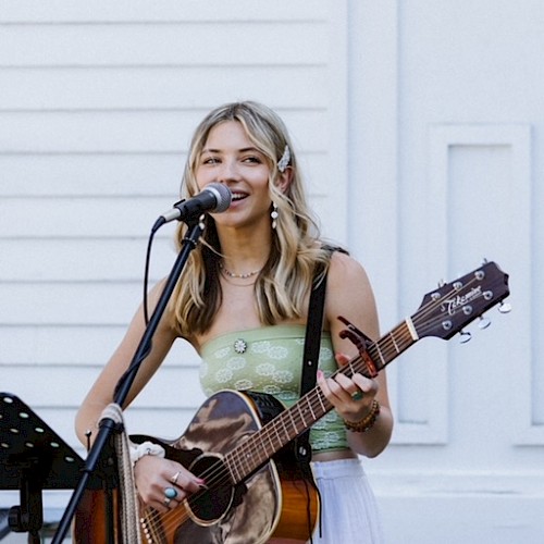 Samantha Hart playing guitar and singing into a microphone outdoors with a music stand and white building in the background, performing live at Currents at Riverhouse Lodge in Bend Oregon on October 26, 2025