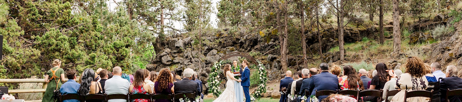 Outdoor wedding ceremony with guests seated on a lawn, a couple standing at the altar, surrounded by trees and greenery, under a cloudy sky.