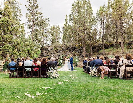 Outdoor wedding ceremony with guests seated on a lawn, a couple standing at the altar, surrounded by trees and greenery, under a cloudy sky.