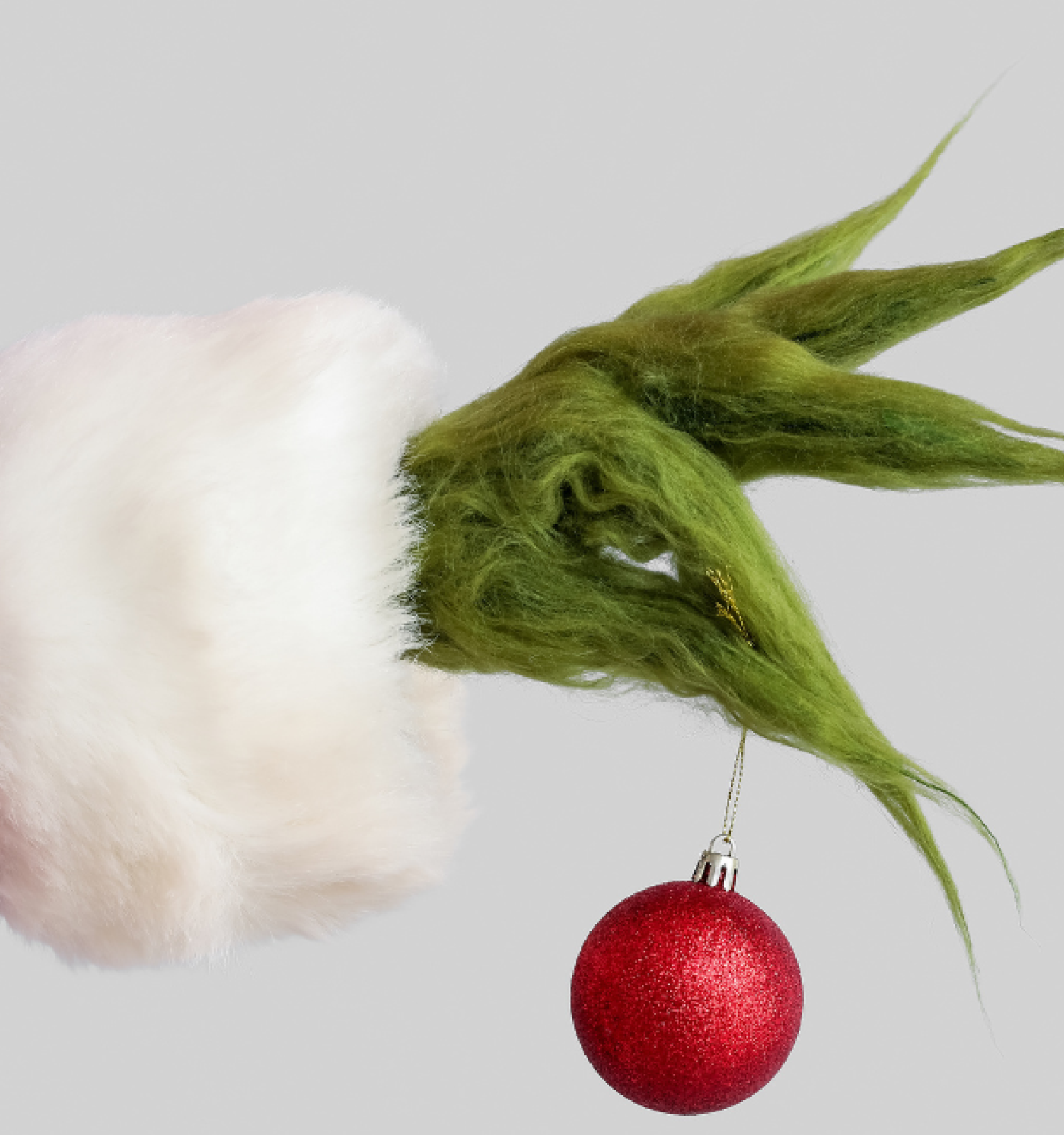 A green, furry hand in a Santa sleeve holds a red Christmas ornament against a gray background.