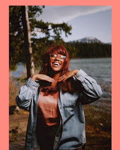 Musician Maria Jackson standing in front of a river with a tree and mountain behind her plays Currents at the Riverhouse November 30th in bend oregon
