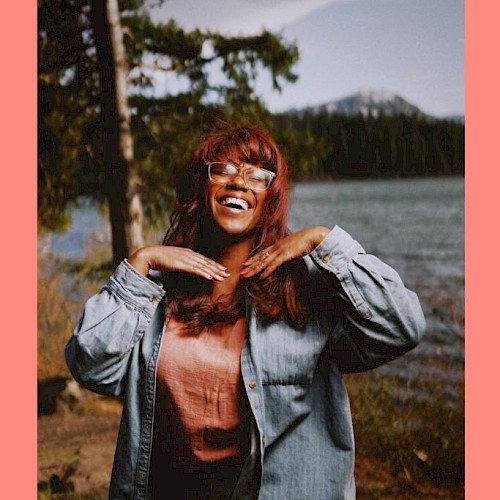 Musician Maria Jackson standing in front of a river with a tree and mountain behind her plays Currents at the Riverhouse November 30th in bend oregon