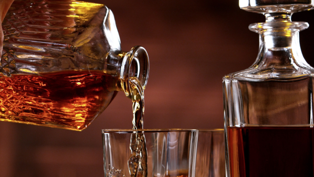 A hand pours whiskey from a decanter into a glass with ice, next to more glasses and a full decanter on a wooden surface.