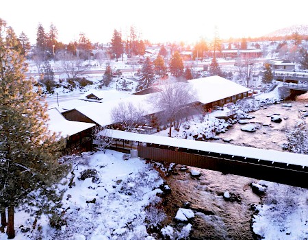 A snowy landscape features a river, a covered bridge, buildings, and trees, with the sun setting in the background.