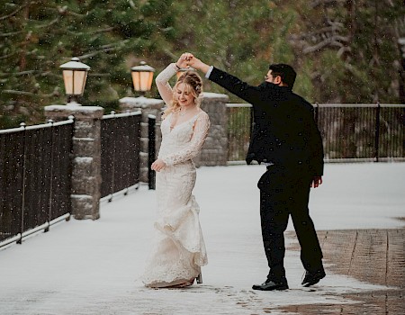 A couple elegantly dances on a snowy path, surrounded by trees and lamps, exuding joy and romance in the wintry setting.