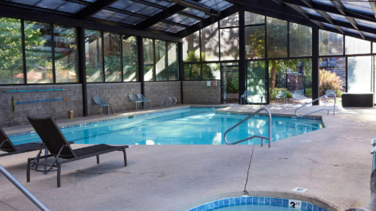The image shows an indoor pool area with a swimming pool, hot tub, lounge chairs, and large windows allowing natural light to enter.