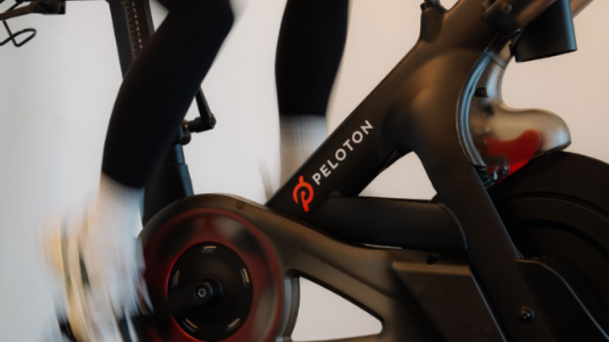 A person is riding a stationary Peloton bike indoors, with motion blur emphasizing the pedaling action.