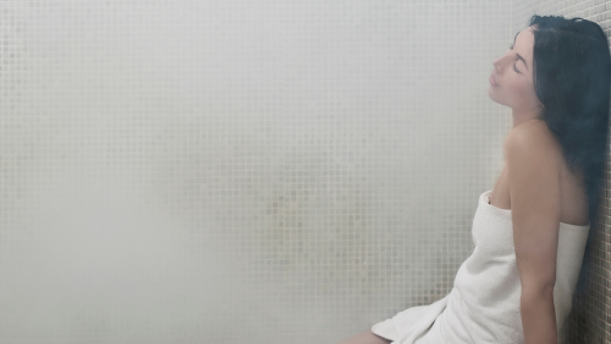 A person wrapped in a towel relaxes in a steamy room, likely a sauna or steam room, with white tiled walls and a serene expression.