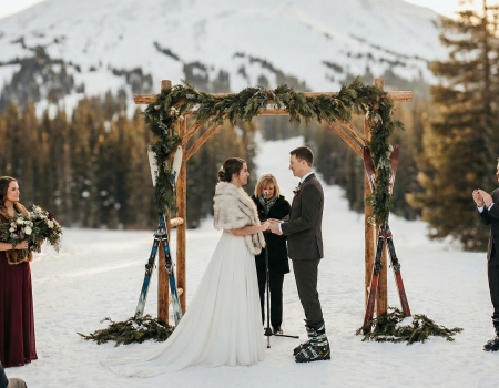 A snowy outdoor wedding under a wooden arch with greenery; couple exchange vows as friends and officiant look on, surrounded by mountains.