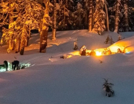 People clearing snow at night with orange portable lights glowing amid snowbanks and trees, a small group gathered around a snow pit.