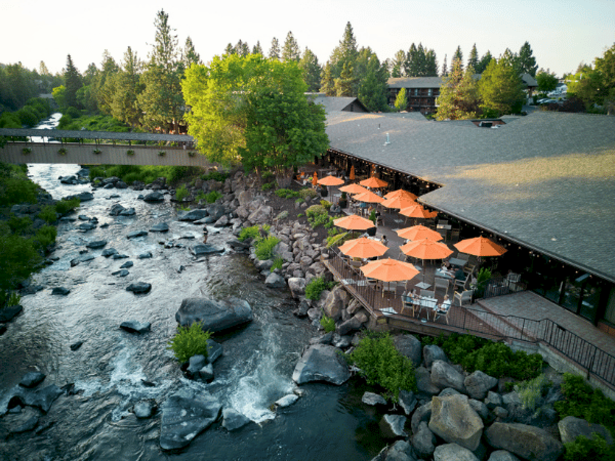 A riverside restaurant with orange umbrellas, stone patio, and rocky shoreline, nestled among trees along a flowing stream.