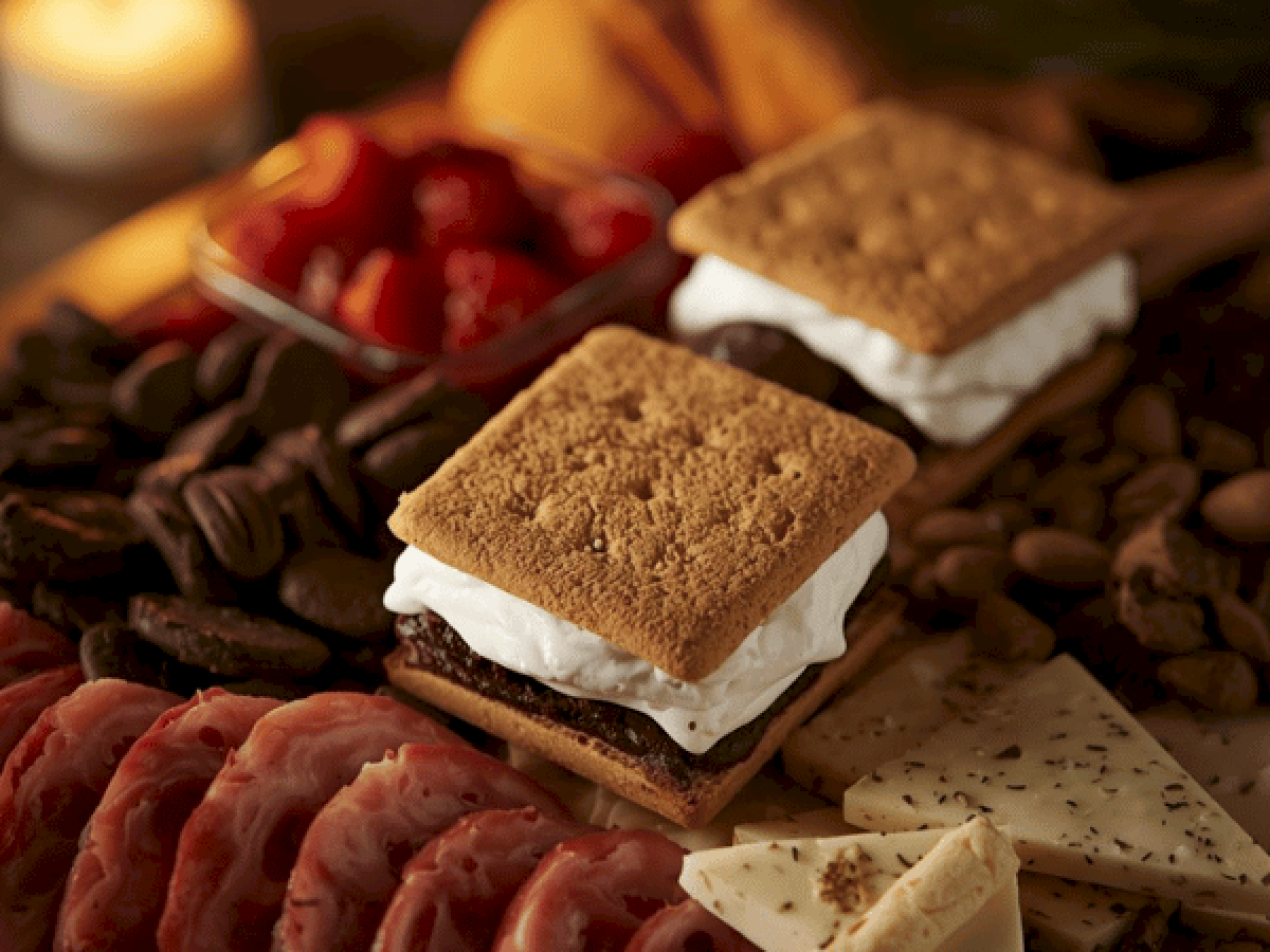 Two graham crackers with marshmallow cream and chocolate, surrounded by chocolate, berries, and cheese on a dessert board.