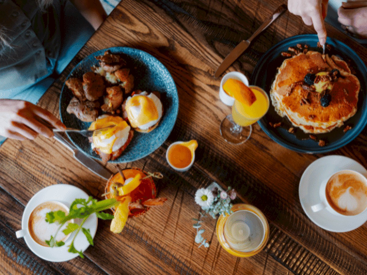 Brunch spread: pancakes, fried eggs, sausages, bacon, toast, a mimosa, orange juice, coffee, and a small salad on a wooden table.