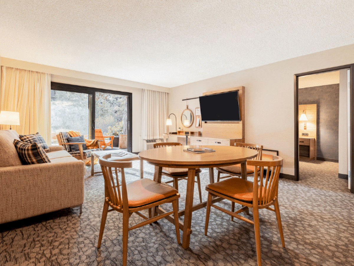 A cozy hotel suite with a round dining table, four wooden chairs, a sofa area, and a wall-mounted TV; sliding glass door opens to a balcony.