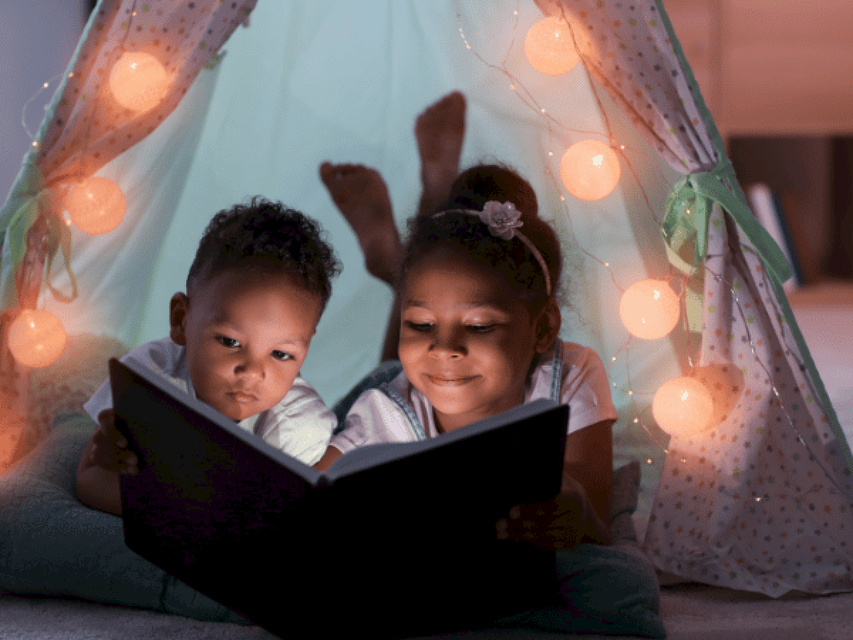 Two kids reading a book inside a cozy, twinkling blanket fort, glowing string lights around them for a magical bedtime scene.
