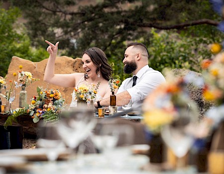 A smiling couple toasting at a decorated outdoor wedding reception, colorful floral arrangements, warm lanterns, and a rustic long table.