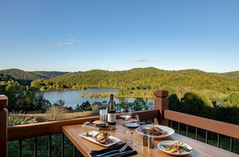 A table with plates of food, wine, and glasses overlooks a scenic lake and lush, green hills on a clear, sunny day.