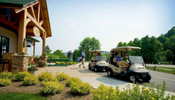 The image shows a golf course scene with two golf carts and a person walking, carrying golf clubs. There is a building and landscaped foliage.