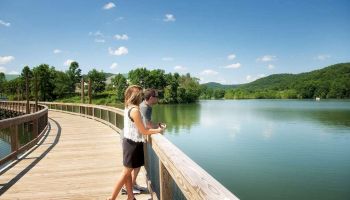 A couple stands on a wooden boardwalk overlooking a serene lake, surrounded by lush green hills under a clear, blue sky with scattered clouds.