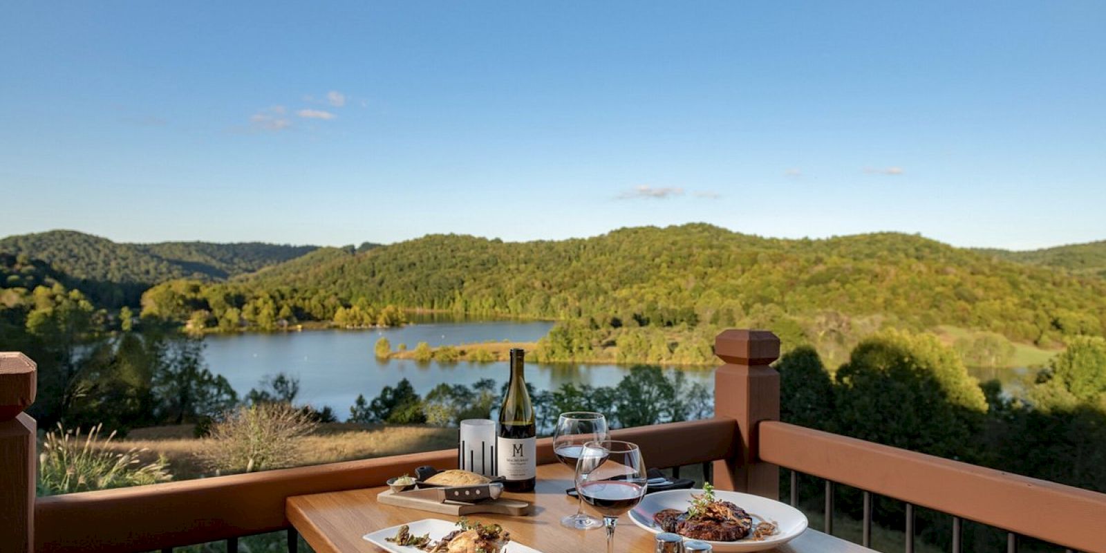 A table set for a meal overlooks a scenic lake with lush green hills. Plates of food, wine, and utensils are ready for dining al fresco.