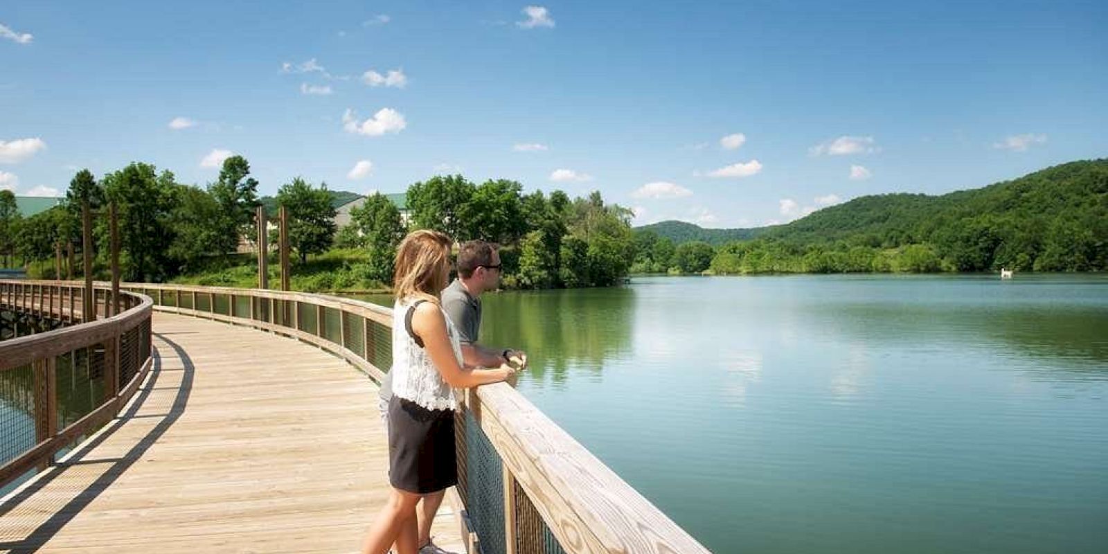 Two people stand on a wooden boardwalk overlooking a serene lake, surrounded by lush greenery and hills under a clear blue sky with scattered clouds.