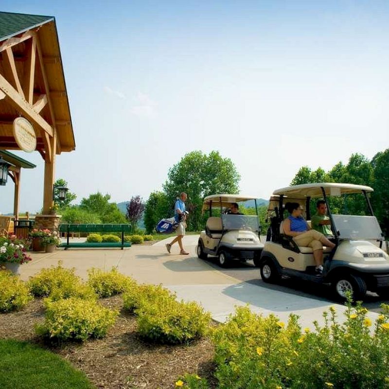 Two golf carts with people sitting in them are parked in front of a building, while another person is walking nearby carrying golf bags.