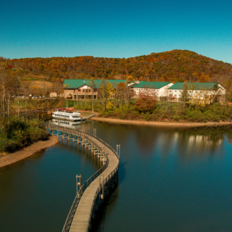 An aerial view of a picturesque lake with a curved boardwalk leading to buildings surrounded by autumn trees and hills under a clear blue sky.