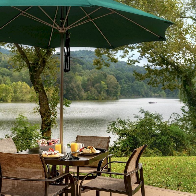 A patio with a table, chairs, and a green umbrella overlooks a serene lake surrounded by trees and hills. It is set for a meal with breakfast items.