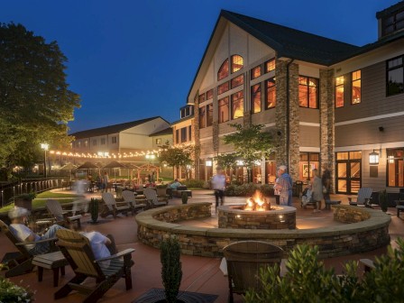 An outdoor seating area at night with a fire pit, string lights, and people relaxing. A large building with lit windows is in the background.