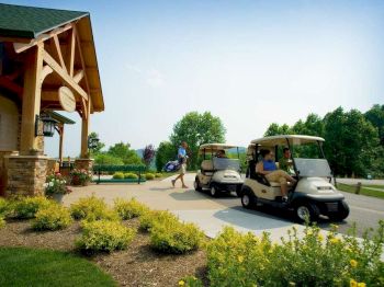 The image shows people near a building, with two golf carts on a paved driveway, surrounded by greenery and flowering plants, suggesting a golf course.