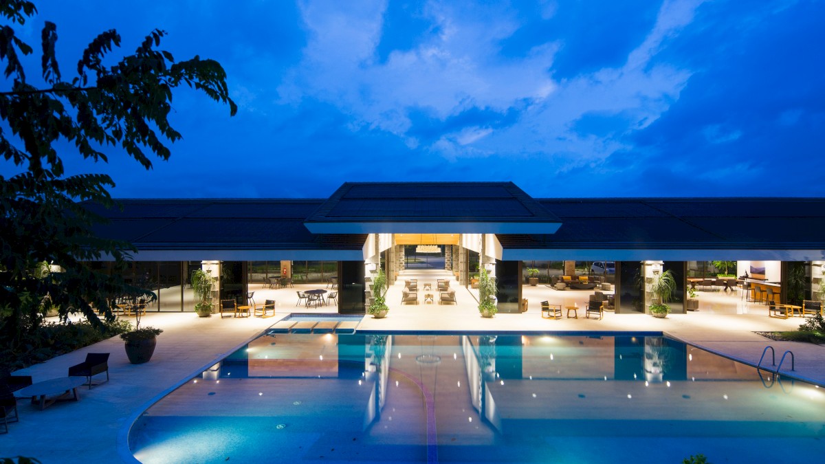 A luxurious evening poolside view of a modern resort with illuminated swimming pool, open-air seating, and sky with scattered clouds.