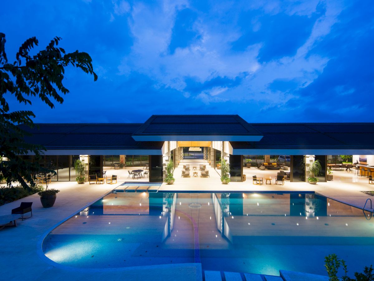 A luxurious evening poolside view of a modern resort with illuminated swimming pool, open-air seating, and sky with scattered clouds.