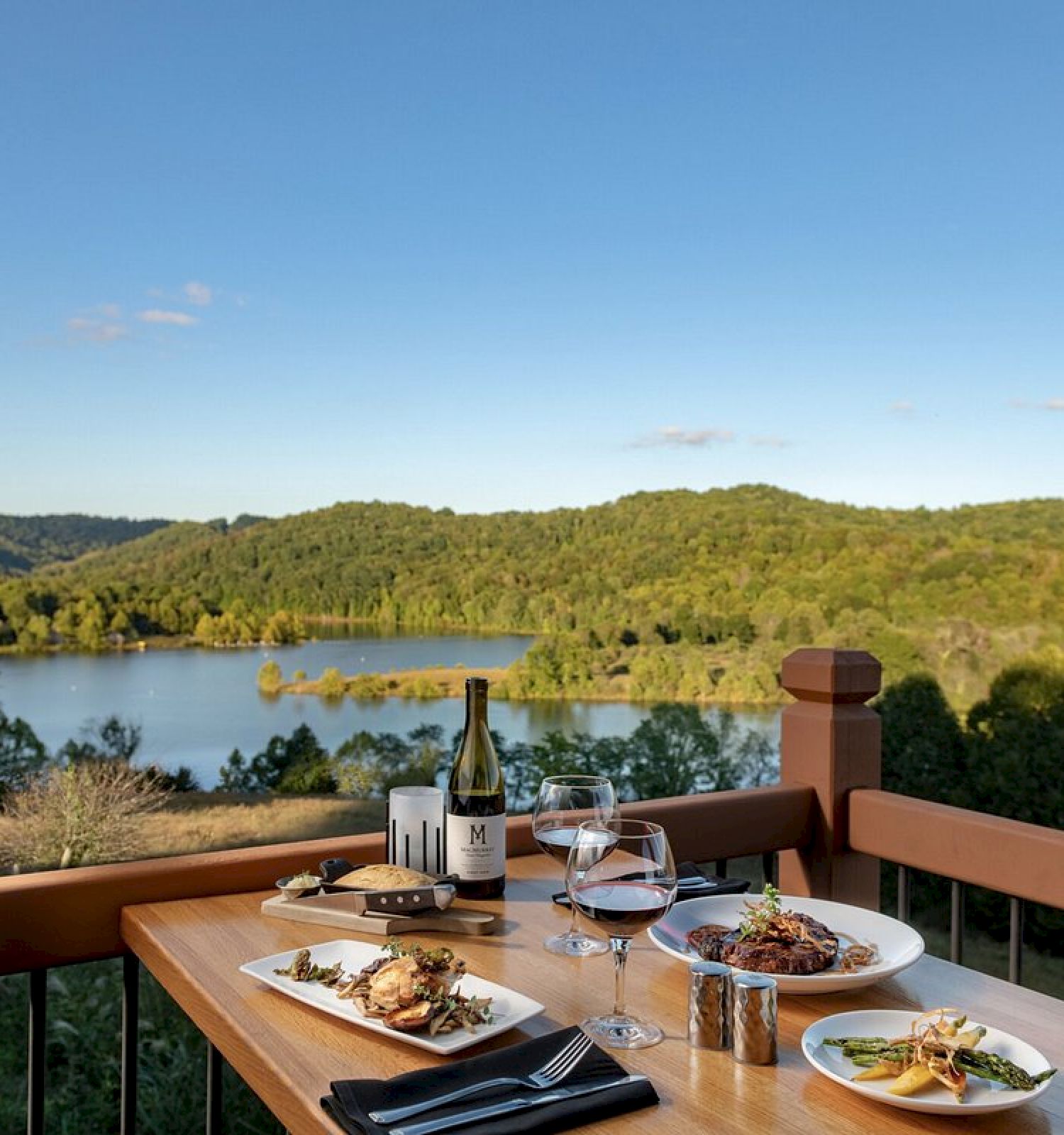 A table set with a meal and wine on a deck overlooking a scenic lake and forested hills under a clear blue sky.