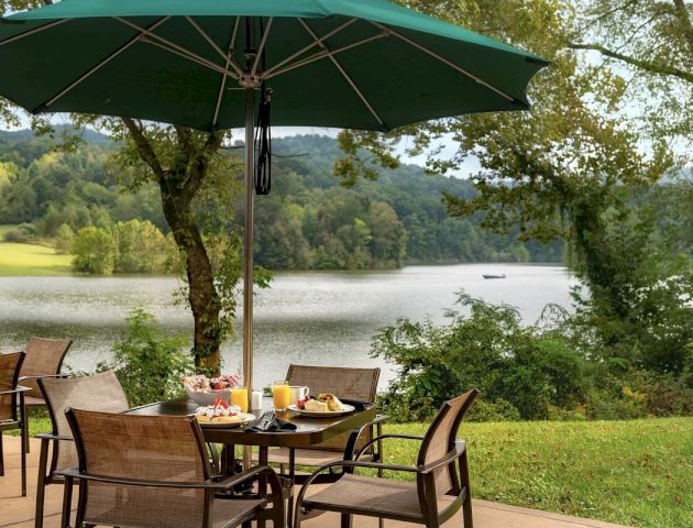 An outdoor dining setup with a green umbrella, chairs, and a table with food and drinks, overlooking a serene lake and lush greenery in the background.