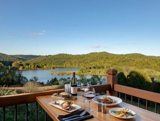 A table set with meals and wine overlooks a scenic lake surrounded by forests and rolling hills under a clear blue sky.