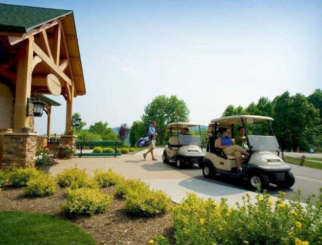 A golf clubhouse with a green roof, yellow flowers in the foreground, and people getting ready to use golf carts in a sunny, green landscape.