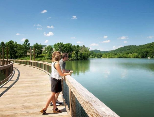 Two people stand on a wooden boardwalk, overlooking a serene lake surrounded by lush greenery and hills under a clear blue sky.