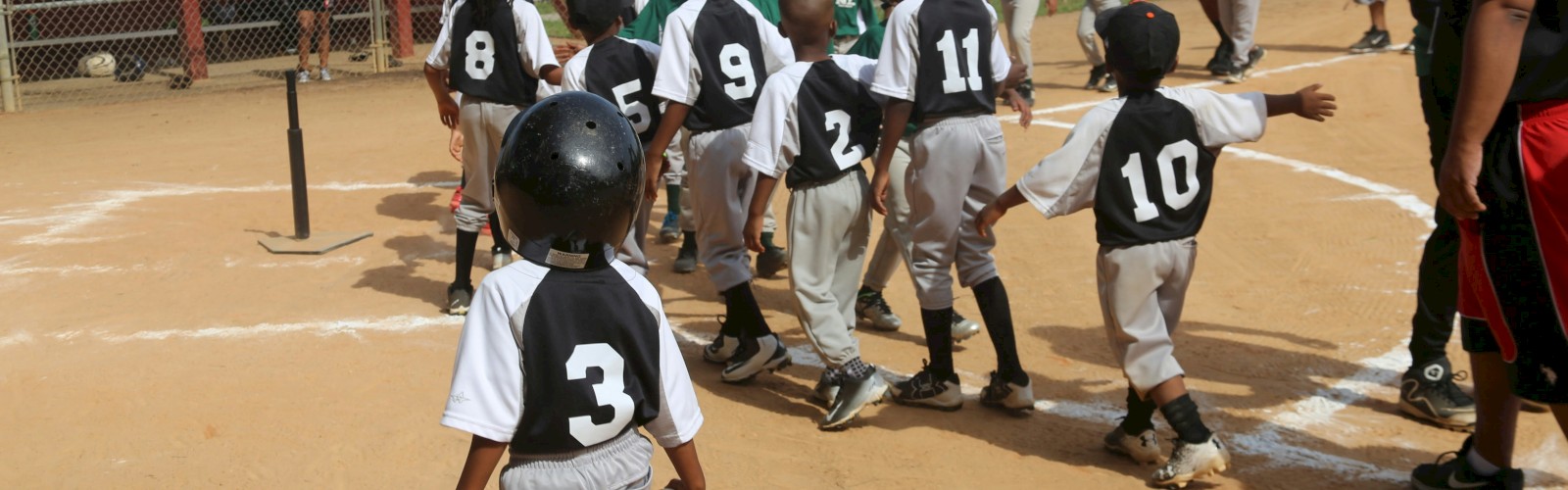 Kids in baseball uniforms walking toward home plate for a group line-up or high-five after a game, on a dusty field.