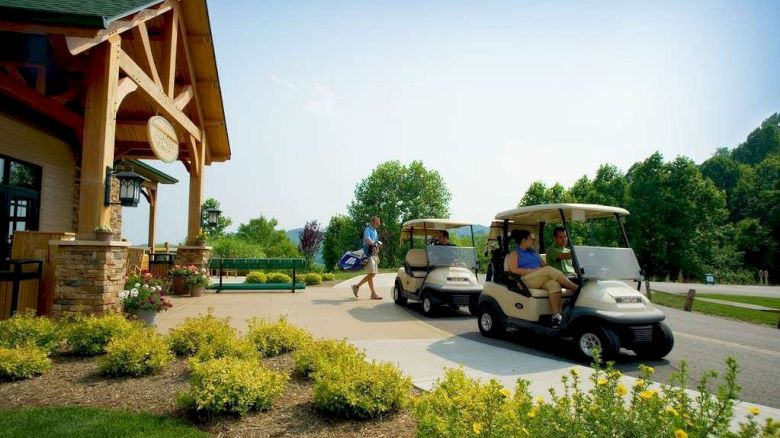 People are at a golf course, with two individuals in golf carts and one person walking towards them carrying a golf bag near a building.