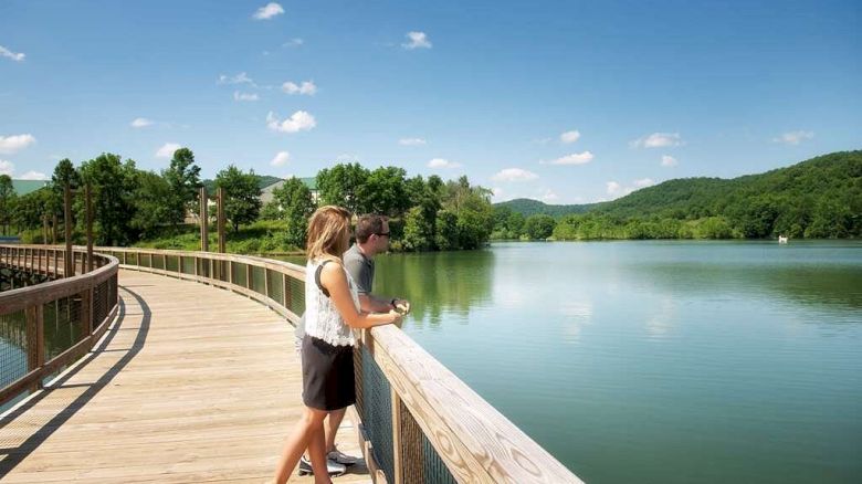 Two people are standing on a wooden walkway overlooking a calm lake with surrounding trees and hills on a sunny day.