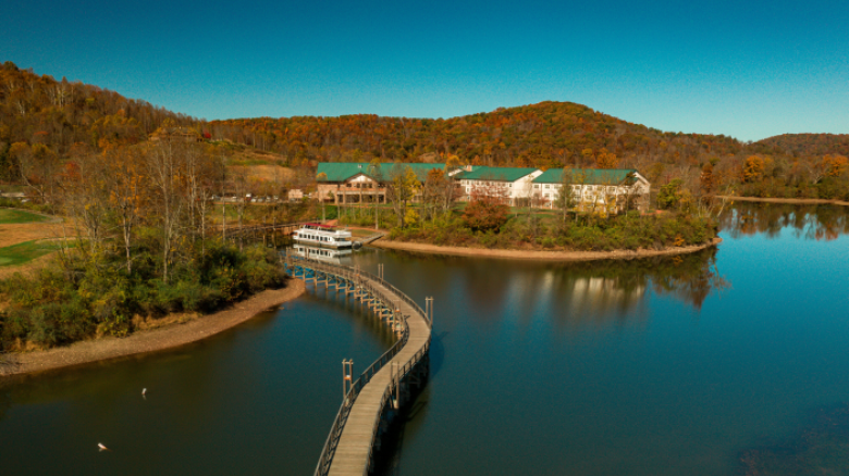 A scenic view of a lake with a curved wooden walkway leading to buildings surrounded by autumn trees and hills under a clear blue sky.