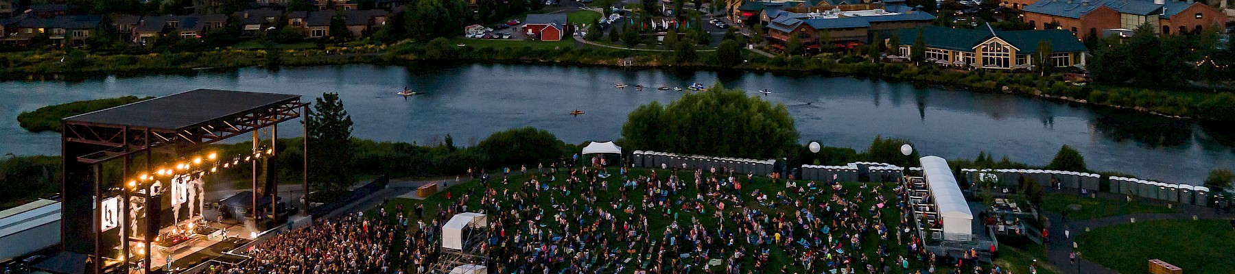 A large outdoor concert by a riverfront, with a stage on the left, a dense crowd of fans, portable tents, and houses across the water in the background.