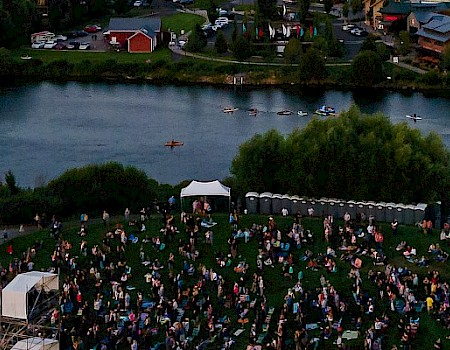 A large outdoor concert by a riverfront, with a stage on the left, a dense crowd of fans, portable tents, and houses across the water in the background.