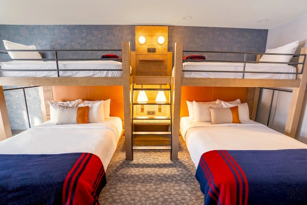 A modern bunk-room with two lower queen beds and two upper twin bunks, a shared center ladder, cozy lighting, and red/blue throws.