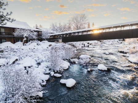 Snow-covered riverfront at Riverhouse Lodge in Bend, Oregon, featuring a footbridge over the Deschutes River at sunrise, highlighting the Wander + Wonder offer with up to 25% off best available rates and a $50 nightly dining credit.