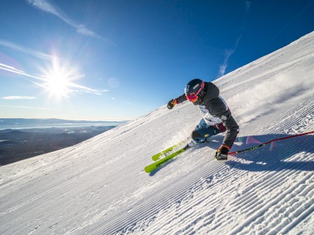 A skier carving a sharp turn down a bright, sunny slope with fresh powder, blue sky, and a blazing sun over the horizon.